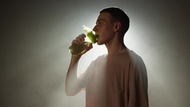 Young man opening bottle and drinking water, standing in dark studio background with white light, side view. Close-up of sportsman having thirsty after workout, drinks from green shaker. 
