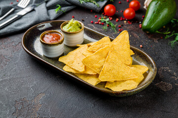 Mexican nachos chips with guacamole sause and tomato sause salsa on brown plate on dark concrete table
