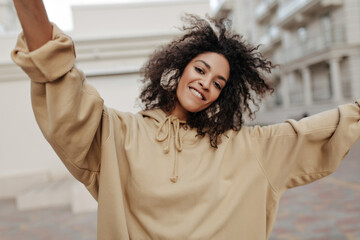 Brunette curly woman in beige hoodie and headphones listens to music outside. Dark-skinned girl on...