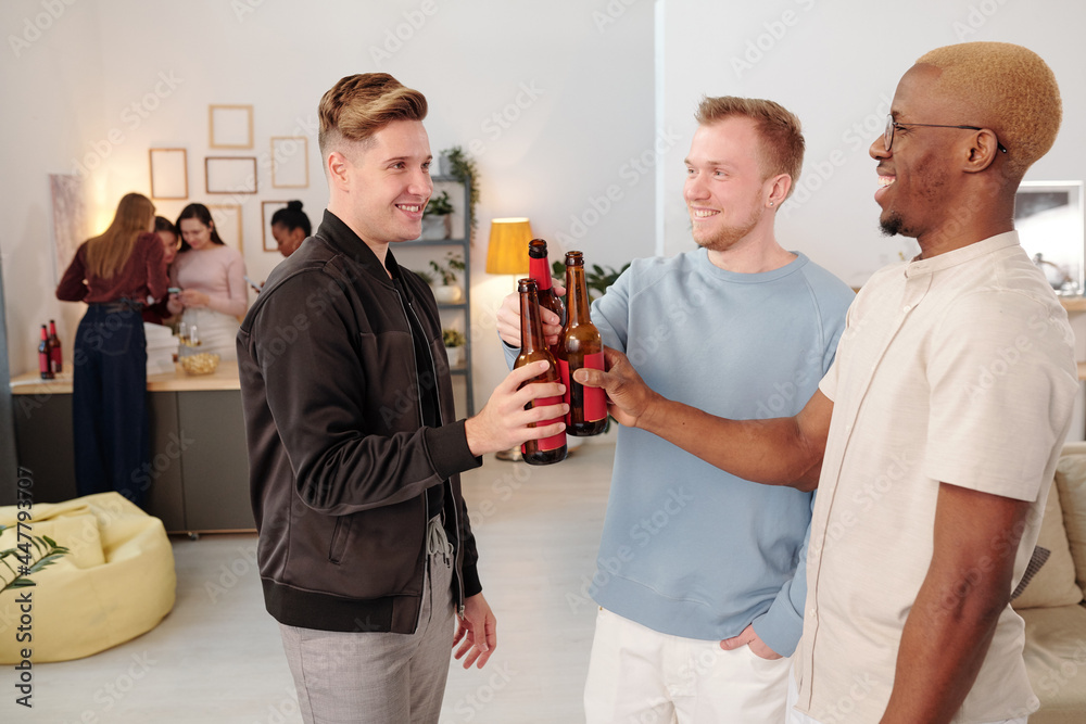 Poster happy intercultural guys clinking with bottles of beer and looking at each other