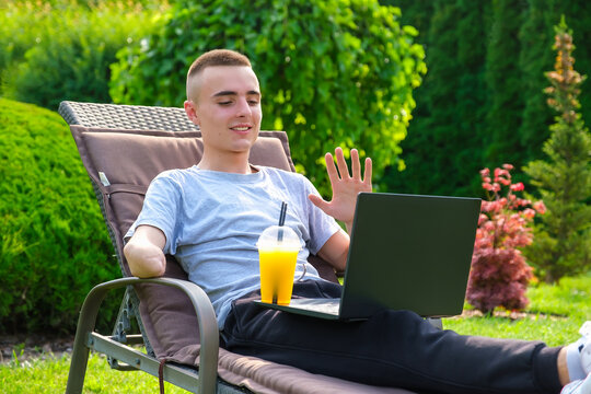 Young smiling guy with amputated arm sits on a sun lounger, drinks a cocktail and talking with friends via social network using a laptop in the yard of his house on the lawn