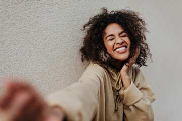 Excited brunette woman in beige hoodie smiles with closed eyes. Dark-skinned girl takes selfie on grey background.