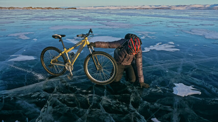 Fototapeta premium Man is riding bicycle on the ice. Ice of the frozen Lake Baikal. Rider is dressed in black down jacket, cycling backpack, helmet. Tires on covered with special spikes. Traveler boy is ride cycle.