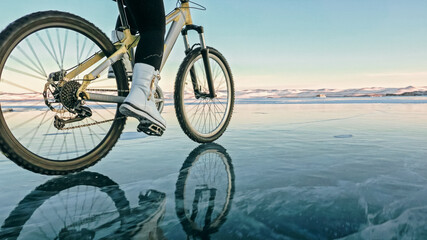 Fototapeta na wymiar Woman is riding bicycle on the ice. Girl is dressed in a silvery down jacket, cycling backpack and helmet. Ice of the frozen Lake Baikal. Tires on bike are covered with spikes. Traveler is ride cycle.