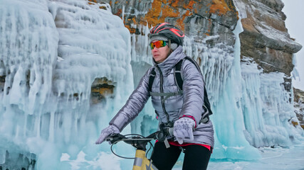 Woman is riding bicycle near the ice grotto. The rock with ice caves and icicles. Girl is dressed in silvery down jacket, cycling backpack and helmet. Tires winter with spikes. Traveler is ride cycle.