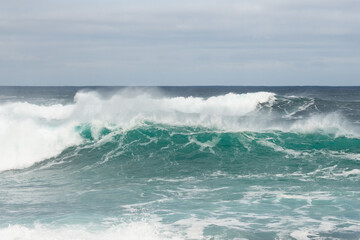 An angry turquoise green color massive rip curl of a wave as it rolls along a beach. The white mist and froth from the wave are foamy and fluffy. The Atlantic Ocean in the background is deep blue. 