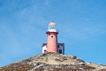 A tall circular red and white lighthouse with an attached light keeper's house. The sky is very dramatic with lots of white clouds and blue sky underneath.  There's grass in front of the lighthouse. 