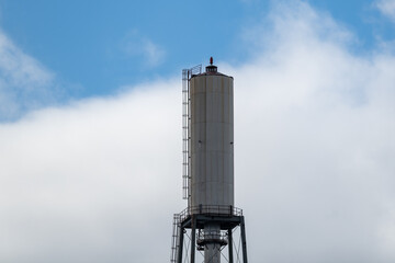 A tall white metal cylinder industrial water tower on four tall legs. There's a ladder on the side of the tower which has a blue sky and white cloudy background. The industrial reservoir is retro.