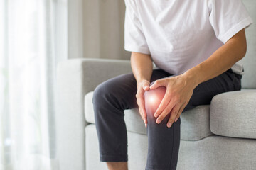 Obraz premium A young man wearing a white T-shirt and gray pants sits on the sofa in the living room at home. He felt pain in his knee area, so he used both hands to hold his knee.