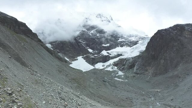 drone view over a desolate receding alpine glacier surrounded by mountains and snow covered rocks and peaks.