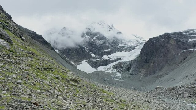 drone view over a desolate receding alpine glacier surrounded by mountains and snow covered rocks and peaks.