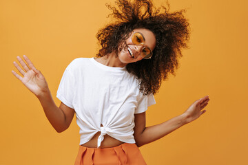 Wonderful woman in white t-shirt and bright sunglasses smiled on isolated backdrop. Dark-skinned girl with curly hair has fun on yellow background..