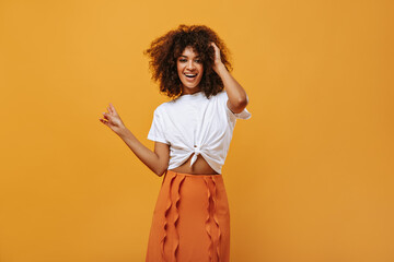 Optimistic lady in white t-shirt and orange skirt smiling on yellow background. Curly girl looks...