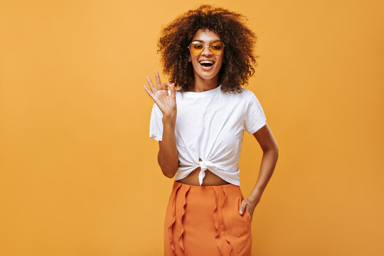 Cheerful girl with fluffy hairstyle in yellow sunglasses mailed on isolated background. Wavy haired woman in white t-shirt posing on bright backdrop..