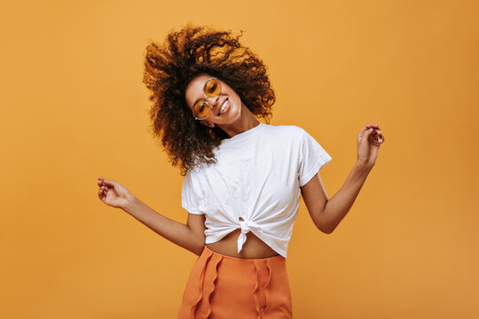 Emotional curly girl in light t-short playing her hair on isolated backdrop. Joyful woman in glasses poses on yellow background..