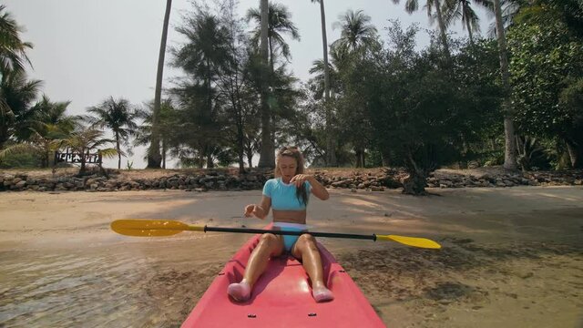 Young woman in sunglasses pulls pink plastic canoe with paddle to azure sea water from beach with palms. The beginning of the boat trip on kayak, boarding sailing. Traveling to tropical countries.