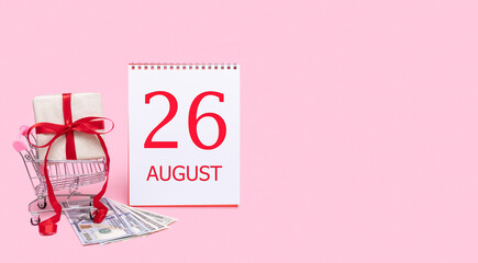 A gift box in a shopping trolley, dollars and a calendar with the date of 26 august on a pink background.