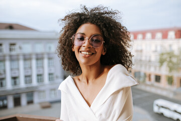 Dark-skinned girl in white clothes and round eyeglasses smiling on balcony. Good-humored woman with...