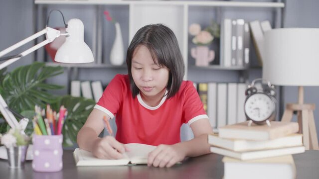 school girl pupil studying at home sitting at desk. Smart cute kid primary school student writing in exercise book thinking homework, learning at table.