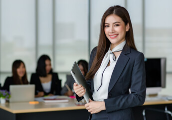 Portrait of young attractive Asian female office worker in formal business suits  smiling at camera in office with blurry colleagues sitting in office as background