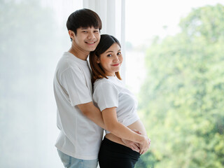 Obraz premium Portrait of a young Asian couple man and pregnant woman dressed casually smiling and standing together by the window with a warm white light from outside. Family concept