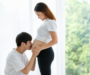 Fototapeta premium portrait of a very lovely young Asian couple. A husband sitting and kissing a wife's belly while a pregnant woman standing smiling and looking at a man with much love