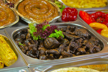 Small mushrooms lie on a large tray next to red bell peppers, various meat sausages and other foods. Delicious food cooked over an open fire, which is offered at a street food fair, event, festival.