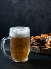 Glass with beer on a dark background. behind it is a wooden board with various beer snacks, crackers, pistachios. Vertical orientation.