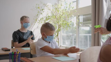 Teacher, children with face mask at school after lockdown, disinfecting hands. school boy holding out his hands while sitting at desk in classroom as female teacher pouring some sanitize - Powered by Adobe