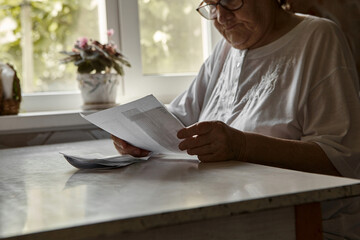 Fototapeta na wymiar Elderly woman sitting at table in kitchen at home holds domestic bills, feels concerned forgot to pay or debt formed. Middle aged 80s retired woman