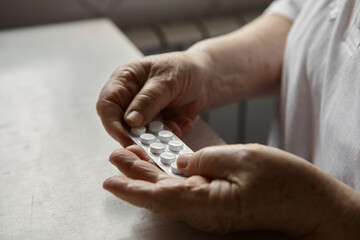 Sad old woman taking pills, health problems in old age, expensive medications. An elderly woman's hands unpacking several pills for taking medication. Grandma takes tablet and drinks a glass of water