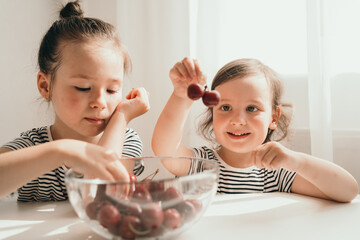 Happy children eat summer berries. Delicious bright juicy berries. Children in striped T-shirts eat. Two sisters eat cherries for an afternoon snack. Children sit at the table and eat