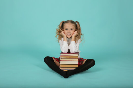 a beautiful blonde schoolgirl girl in glasses with red frames with ponytails in a white shirt a red plaid skirt with black socks and shoes is sitting in the background and put her hands on a stack of