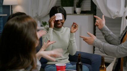 Multiracial friends playing funny game using sticky notes having fun together late at night sitting on couch in living room. Group of multi-ethnic people drinking beer, eating chips enjoying free time