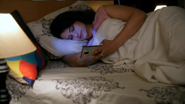 Portrait of a pretty woman using her smartphone lying down on the bed at night. Beautiful Indian lady in casual clothes - modern lifestyle  late night calls  mobile addiction 