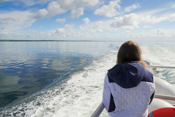 Fototapeta premium Teenager girl on a ocean voyage watching ship trail and ocean scenery. Galway bay, Ireland. Travel and tourism concept. Blue cloudy sky. The model in white jacket, has long hair