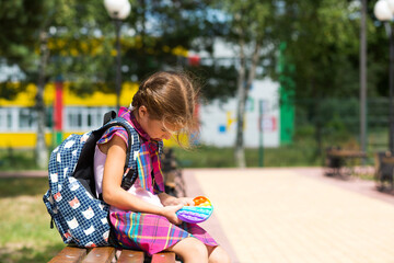 Little girl with a backpack and in a school uniform in the school yard plays pop it toy. Back to school, September 1. The pupil relaxes after lessons. Primary education, elementary class for student