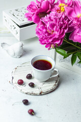 Shabby chic tea. A cup of tea on a white wooden tray with a cherry nearby. Peonies are in a white box. Blurred background