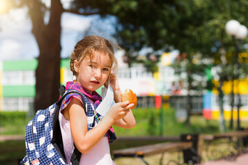 Girl with a backpack take off the medical mask and eating pie near the school. A quick snack with a bun, unhealthy food, lunch from home. Back to school. Education, primary school classes, September 1