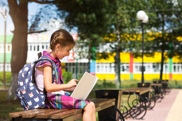 Girl with a backpack sitting on a bench and reading a book near the school. Back to school, lesson...