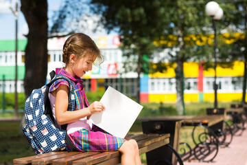 Girl with a backpack sitting on a bench and reading a book near the school. Back to school, lesson schedule, a diary with grades. Education, primary school classes, September 1