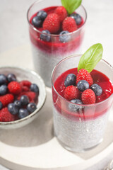 Two glasses with chia seeds pudding, raspberry jam and fresh raspberries and blueberries on a concrete tray on grey surface, a close up