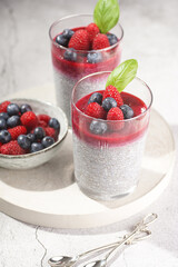 Two glasses with chia seeds pudding, raspberry jam and fresh raspberries and blueberries on a concrete tray on grey surface, a close up