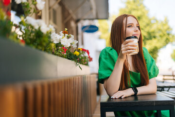 Portrait of attractive young woman enjoying tasty cup of coffee sitting at table in outdoor cafe terrace in summer day. Pretty redhead lady relaxing leisure activity on urban street.