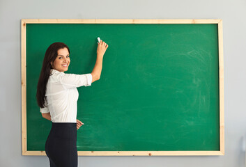 Female teacher looking at camera and writing something on chalkboard. Happy tutor giving online...