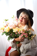 Happy beautiful woman with long blond hair and in black hat posing with flowers in studio on white background