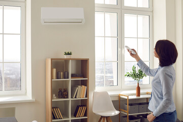 Woman switches wall air conditioner at home. Young lady standing in her modern light living room...