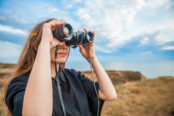 young woman on a high hill by the sea looking through binoculars in summer