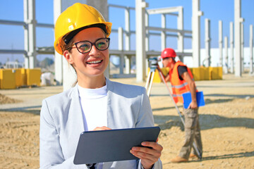 Happy beautiful female architect with tablet on construction site. She is smiling and satisfied with her job, behind her construction engineer with measuring device, positive emotions