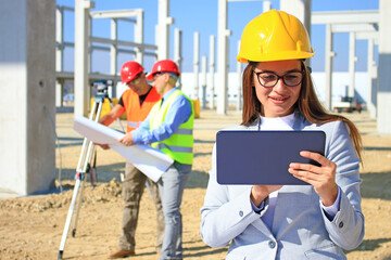 Happy beautiful female architect with tablet on construction site. She is smiling and satisfied with her job, behind her construction engineers with measuring device, positive emotions